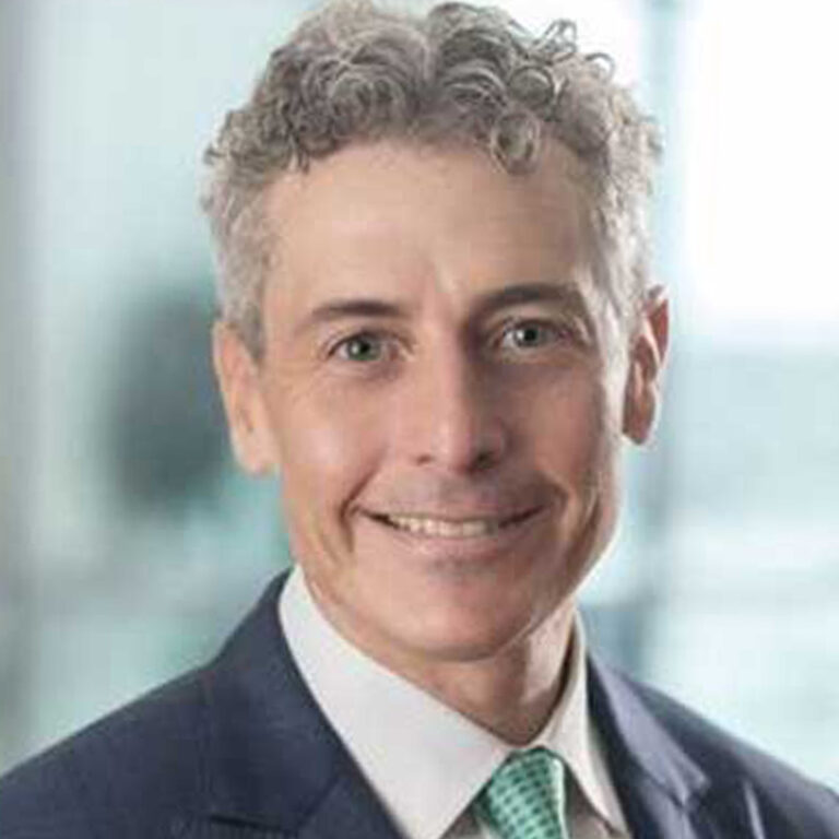 Smiling man with short curly gray hair wearing a dark suit jacket, white shirt, and light green tie, set against a blurred indoor background.