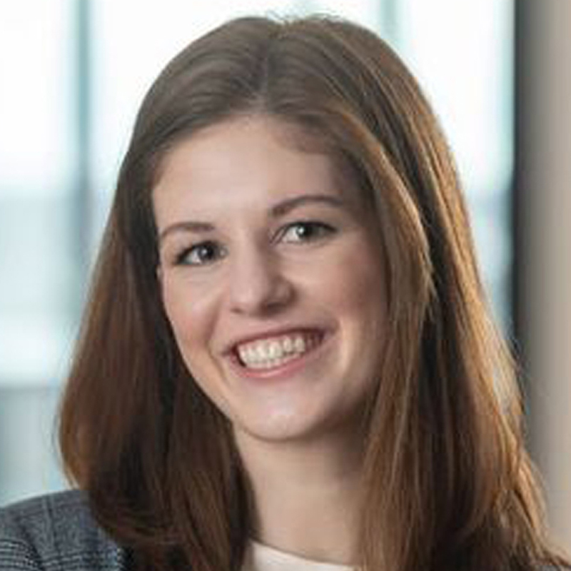 A young woman with long brown hair, smiling, wearing a gray blazer and white top, posed in front of a softly blurred indoor background.