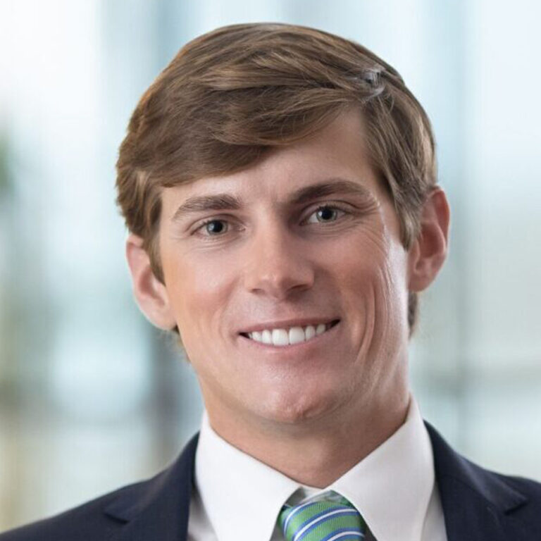 A smiling man with short brown hair wearing a dark suit, white shirt, and a green striped tie, standing in front of a blurred light background.