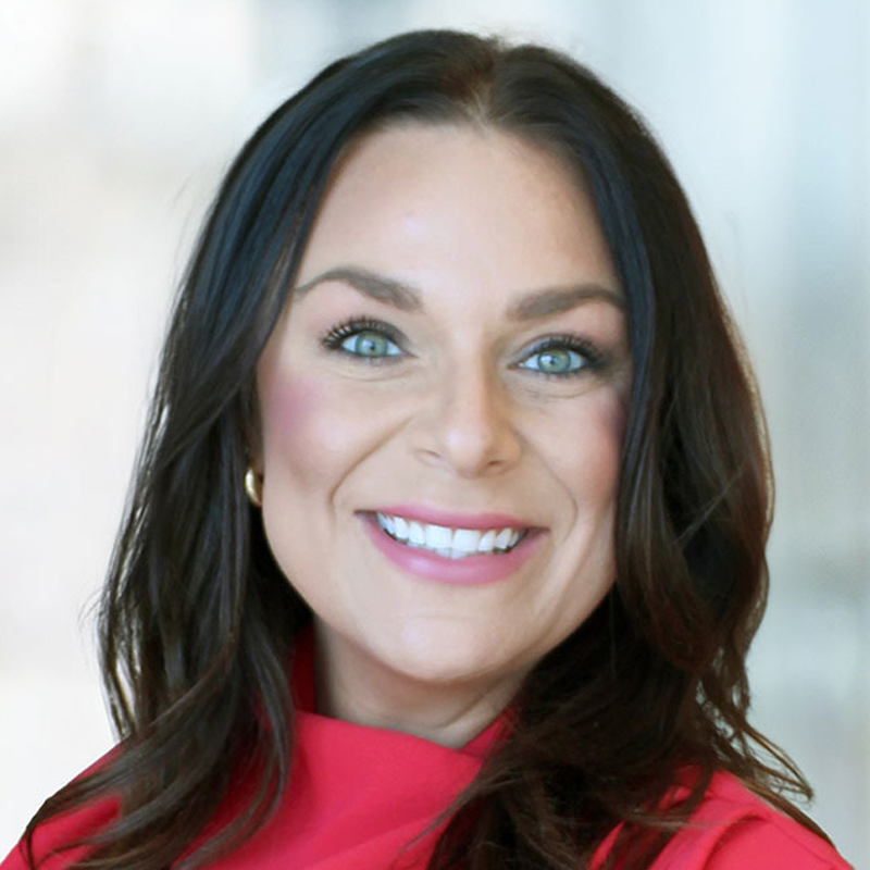 A woman with long dark hair and blue eyes smiles at the camera. She is wearing a bright pink top and gold hoop earrings, with a blurred light background.