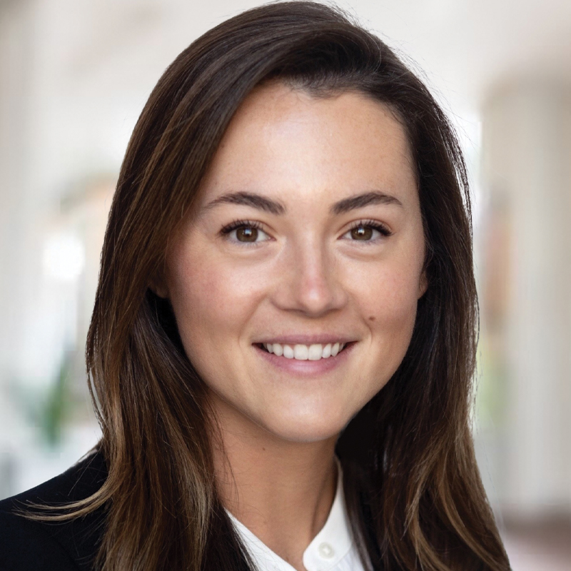 A woman with long brown hair, brown eyes, and a bright smile, wearing a black blazer and white shirt, stands in a softly blurred indoor setting.