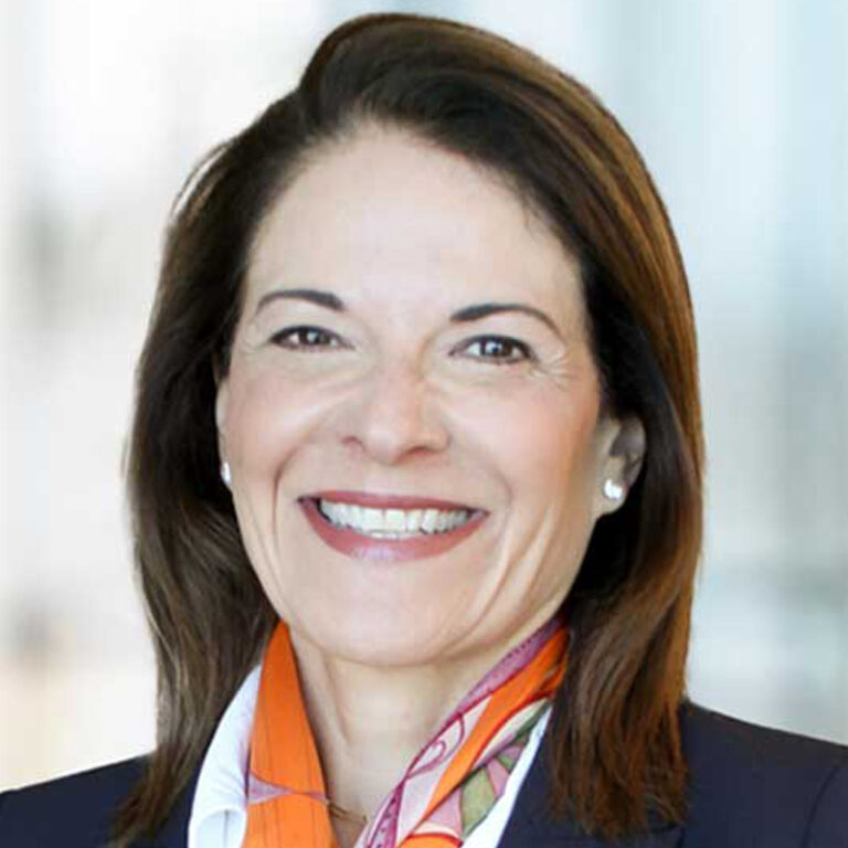 A smiling woman with straight brown hair, wearing a dark blazer, white blouse, and an orange patterned scarf, poses in front of a blurred light background.