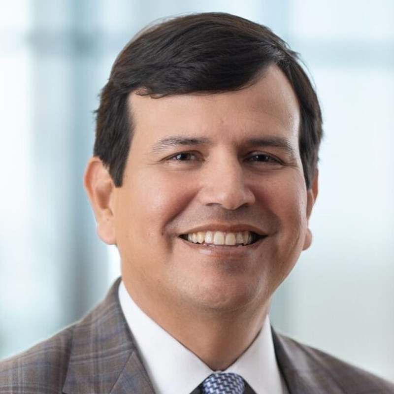 A man with dark hair in a gray plaid suit, white shirt, and patterned tie smiles at the camera. The background is softly blurred with shades of blue and white.