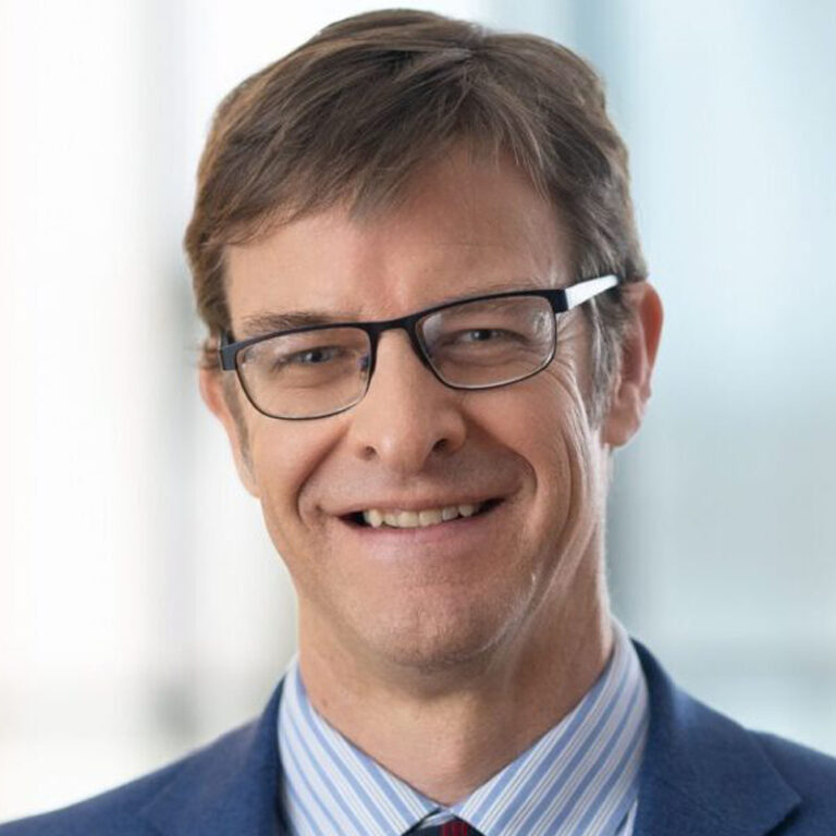 A middle-aged man with short brown hair and glasses is smiling. He is wearing a blue suit, striped shirt, and tie, with a blurred light background.
