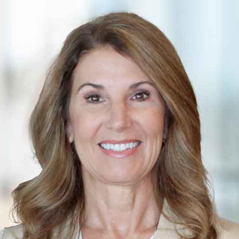 A woman with long, wavy light brown hair smiles at the camera. She is wearing a light-colored blazer and is posed in front of a softly blurred background.