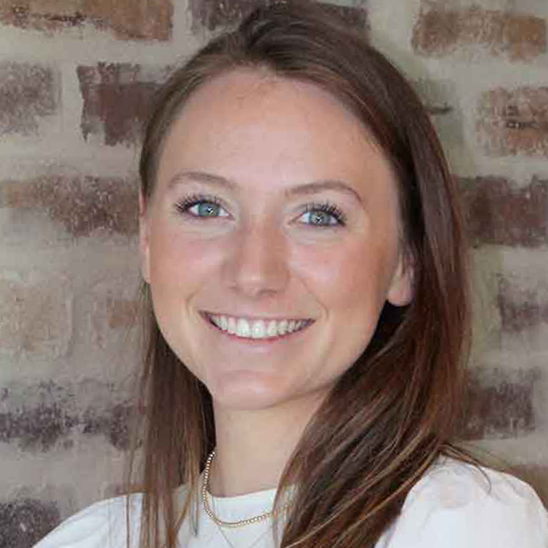 A young woman with long brown hair smiles at the camera. She is wearing a white top and a gold necklace, standing in front of a brick wall.