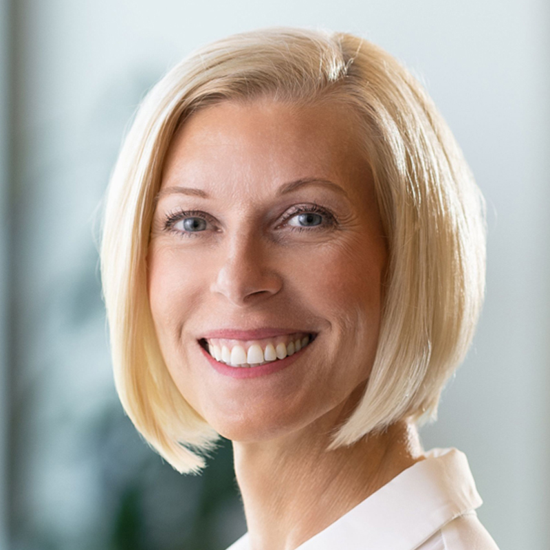 A woman with straight, chin-length blonde hair and blue eyes smiles warmly at the camera. She is wearing a white top and the background is softly blurred with hints of green.