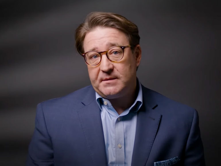Professional headshot of Bryan Hutto wearing a navy blazer and glasses, set against a dark studio background.