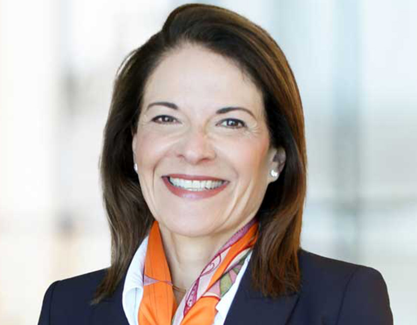 Melissa Anderson wearing a dark blazer, a white shirt, and an orange patterned scarf. The background is softly blurred with light colors.
