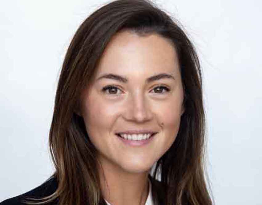 Mary Mullins Romanczuk wearing a dark blazer over a white top, and the background is plain and light-colored.