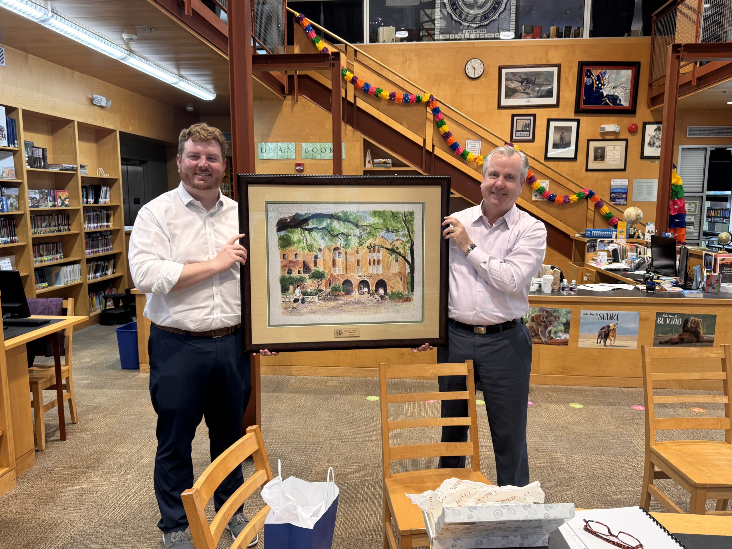 Two men standing in a library hold up a framed painting of a building and trees. The library shelves, artwork, and colorful decorations are visible in the background.