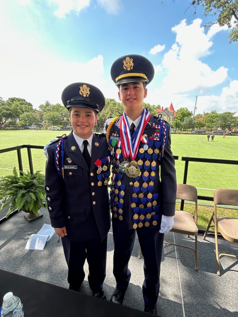 Two young people in decorated military uniforms stand smiling side by side outdoors, in front of an open grassy field. One wears many medals and ribbons; the other has a few medals. Its a bright, sunny day.