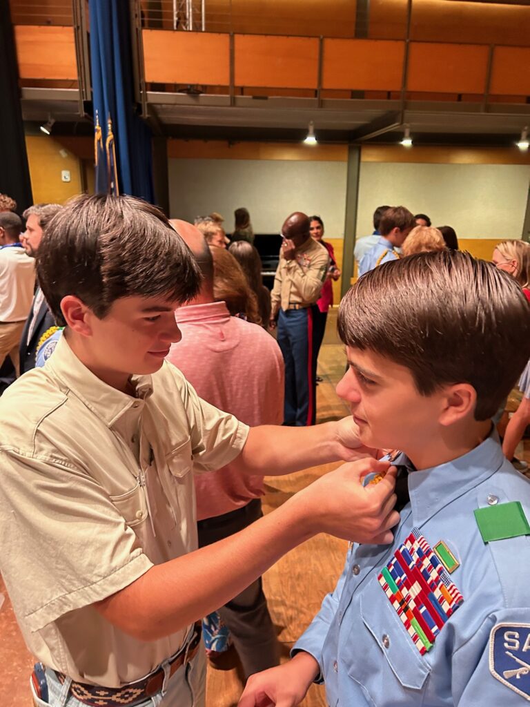 Two boys stand indoors among a group; one in a tan shirt helps the other, who wears a blue uniform covered in colorful badges and ribbons, adjust something on his collar. Other people are gathered and talking in the background.
