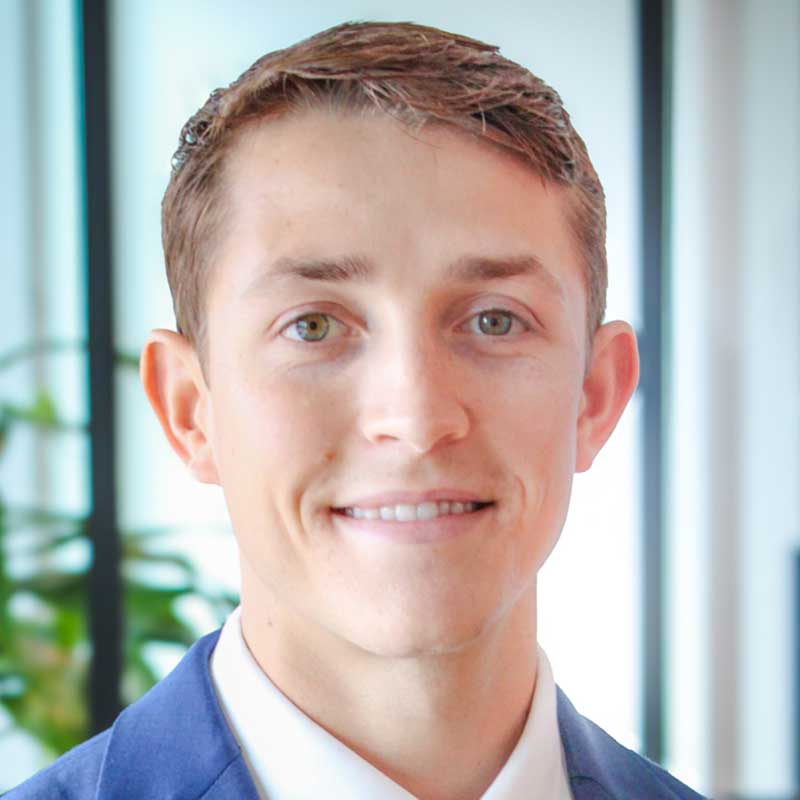 A young man with short brown hair, wearing a blue suit and white shirt, smiles at the camera. The background is bright with blurred green plants and window light.
