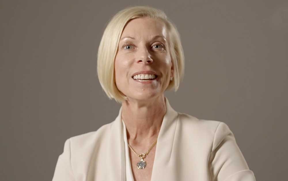 A smiling woman with short blonde hair, wearing a light-colored blazer and a gold necklace, stands against a plain gray background.