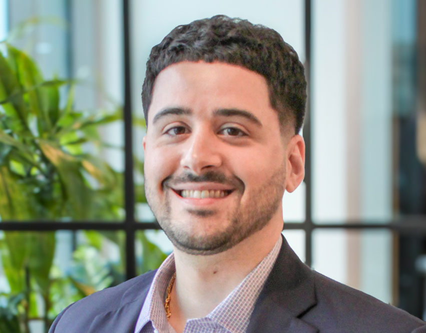 A man with short curly hair and a trimmed beard, wearing a dark blazer and light shirt, smiles at the camera. Blurred green plants and large windows are in the background.