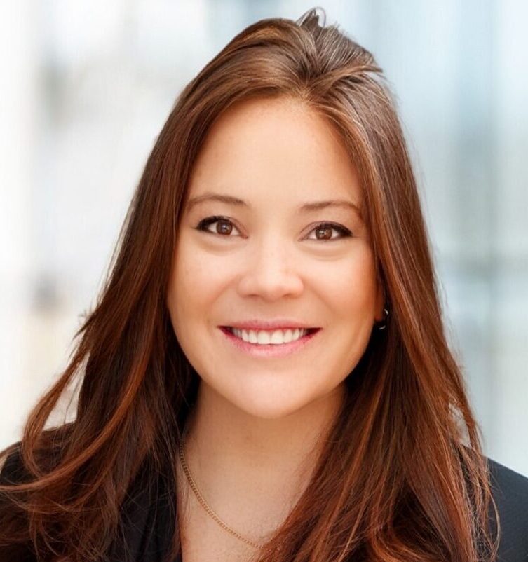 A woman with long brown hair, wearing a black blazer, black top, and beige pants, stands indoors with hands in her pockets, smiling at the camera. The background is softly blurred, showing a modern, bright interior.