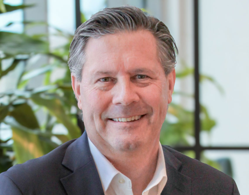 A middle-aged man with short, grayish-brown hair, wearing a dark suit jacket and white shirt, smiling in front of a blurred background featuring green plants and large windows.