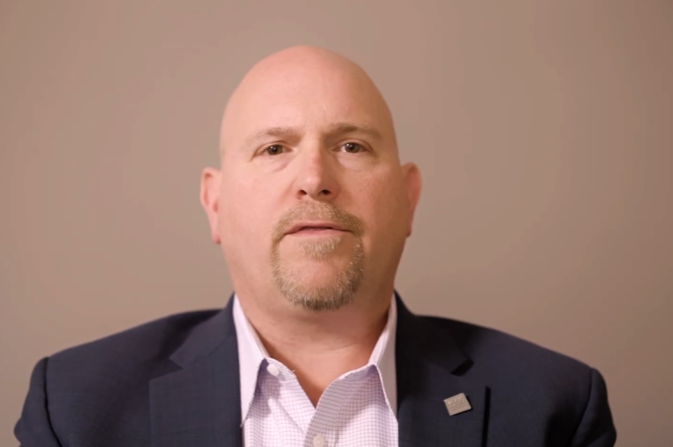A man in a suit and pink shirt sits in front of a plain beige background. 1900 Wealth. is written in white text at the top right corner of the image.