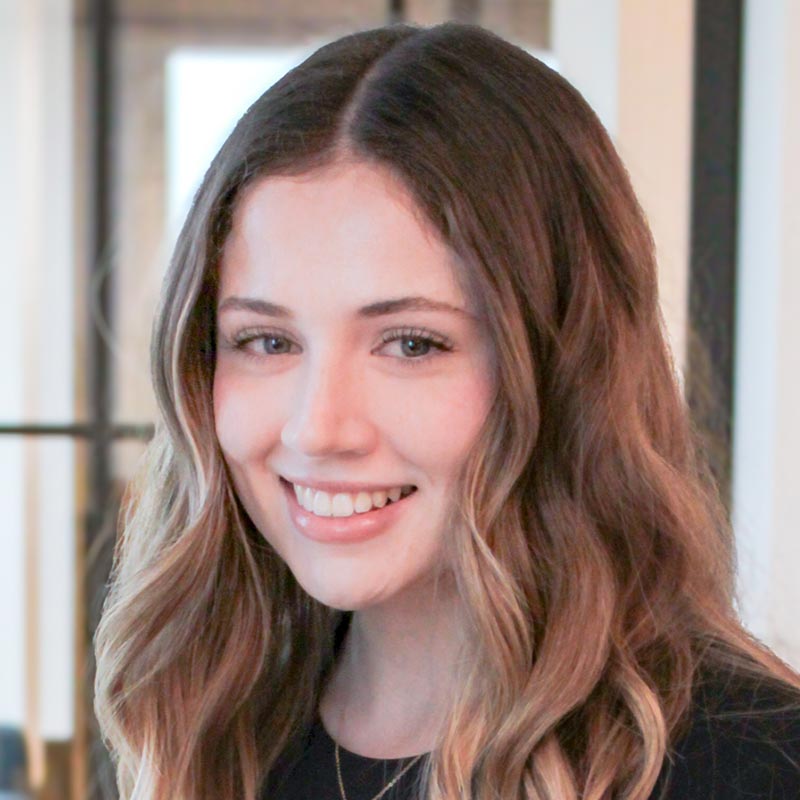 A young woman with long, wavy brown hair smiles at the camera indoors. She is wearing a black top and standing in a well-lit, modern setting with blurred background elements.